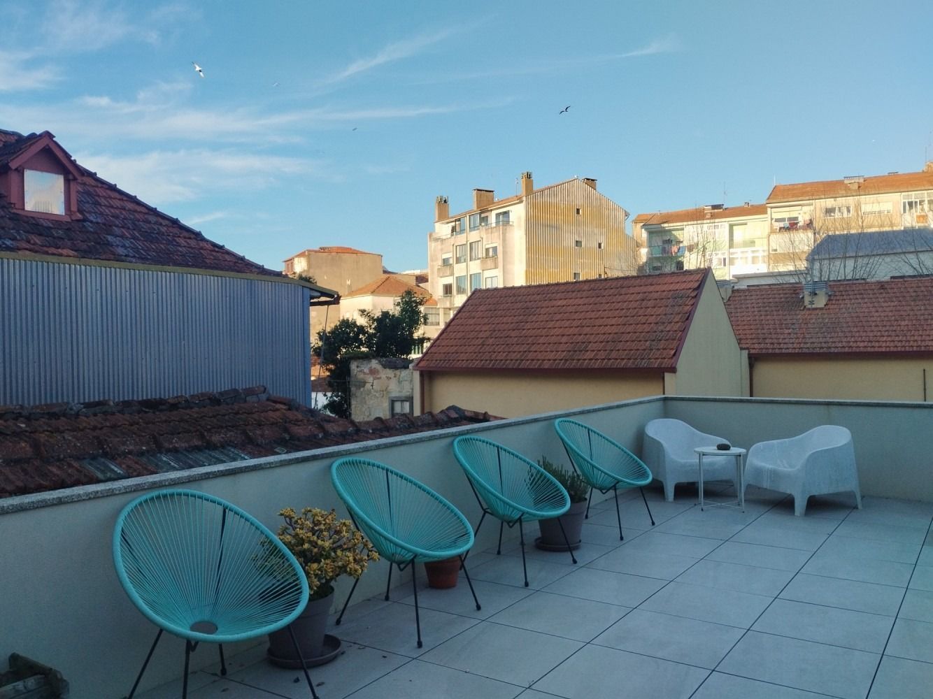 Rooftop terrace with turquoise chairs, white tables, and city buildings under a blue sky