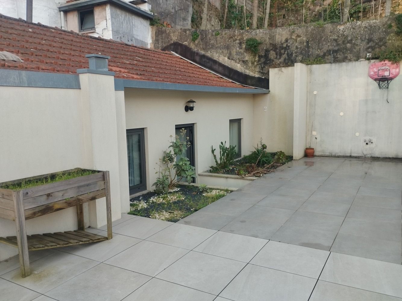 Empty tiled courtyard beside a cream house with a red-tiled roof and a small planter bench.
