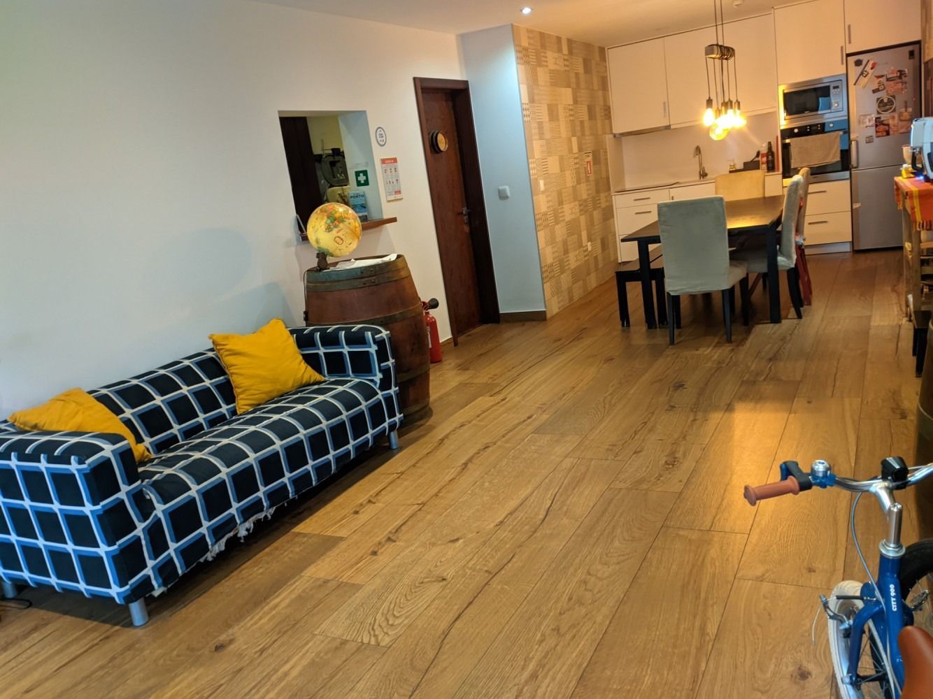 Living room with blue plaid sofa, wooden floor, dining table, and kitchen in the background