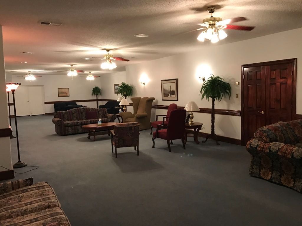 Spacious waiting area with couches, chairs, tables, and plants. Ceiling fans and wall sconces illuminate the room.
