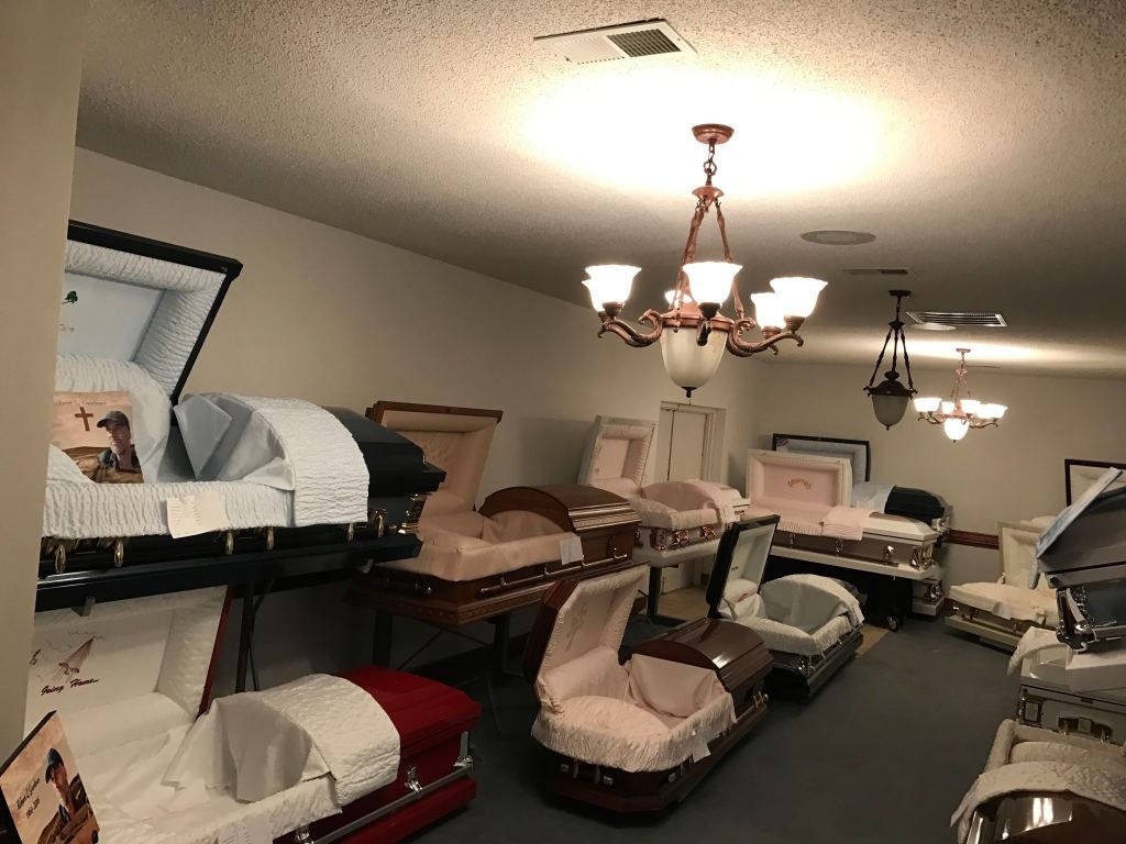 Interior of a funeral home displaying various open coffins; a chandelier hangs from the ceiling.