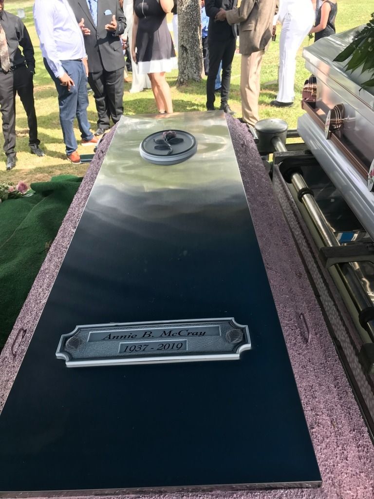 Coffin at a funeral, with nameplate and a logo. People in the background are standing on grass.