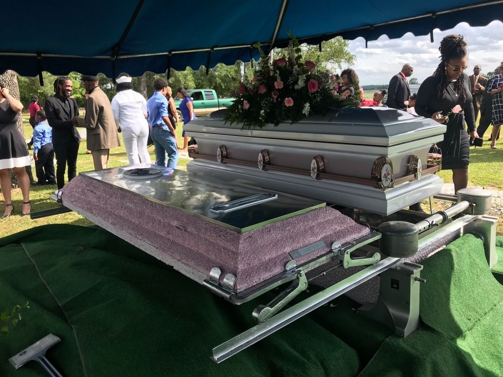 Two open caskets on a grassy area, people gathered nearby under a canopy.
