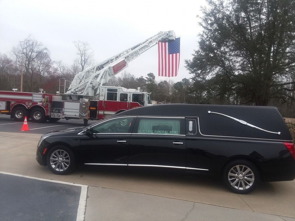 Black hearse with a fire truck ladder displaying an American flag.
