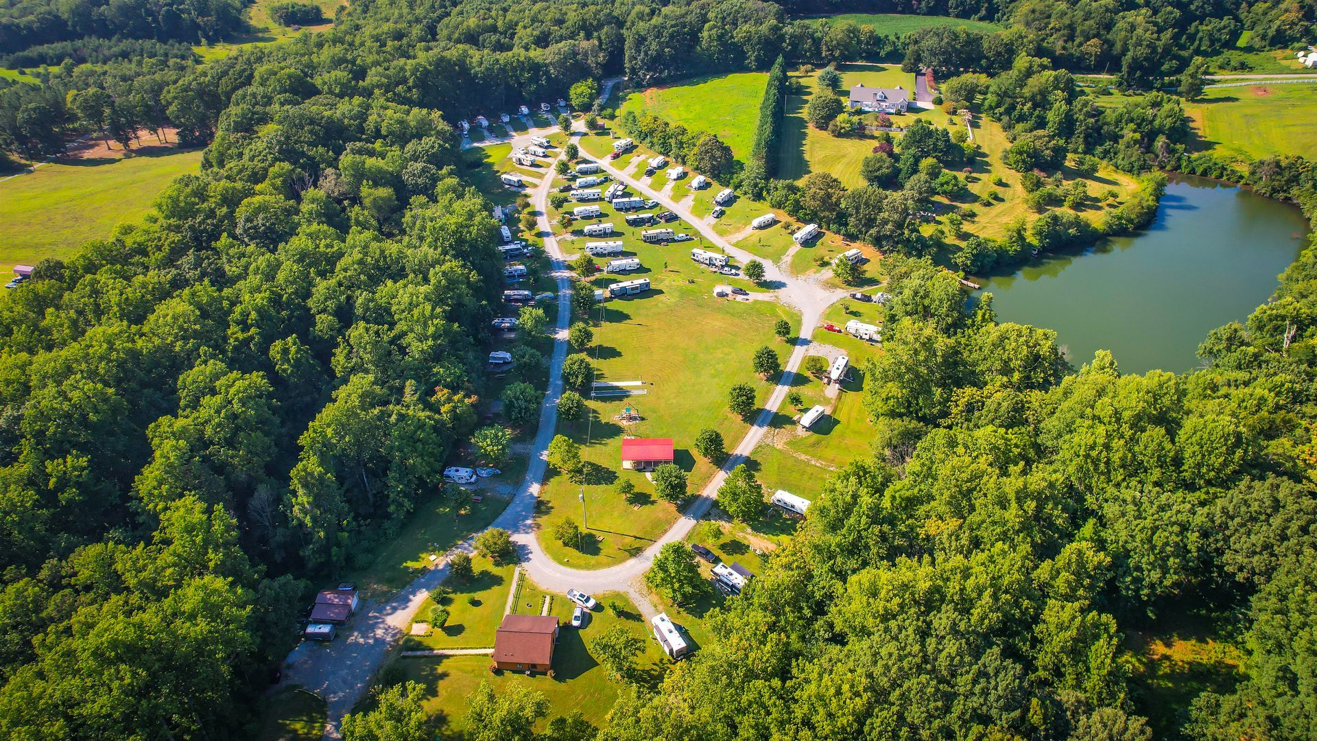 Aerial view of a campground with RVs, surrounded by trees, a lake, and fields under a sunny sky.
