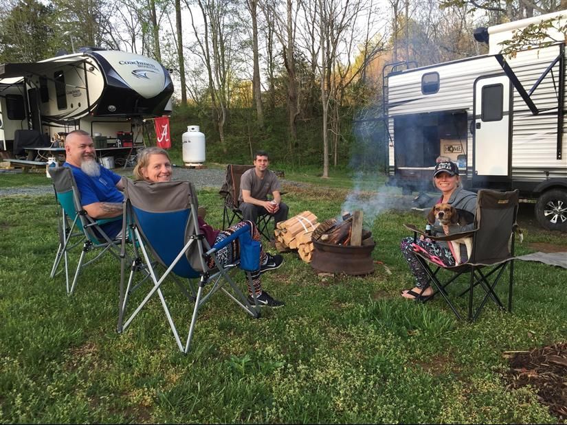 Four people relax around a campfire at a campsite, with RVs in the background.