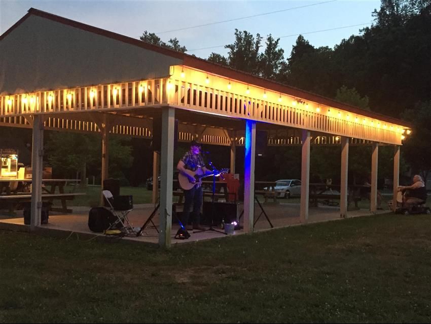 Man playing guitar under a lit pavilion at dusk. Picnic tables, trees, and cars in background.