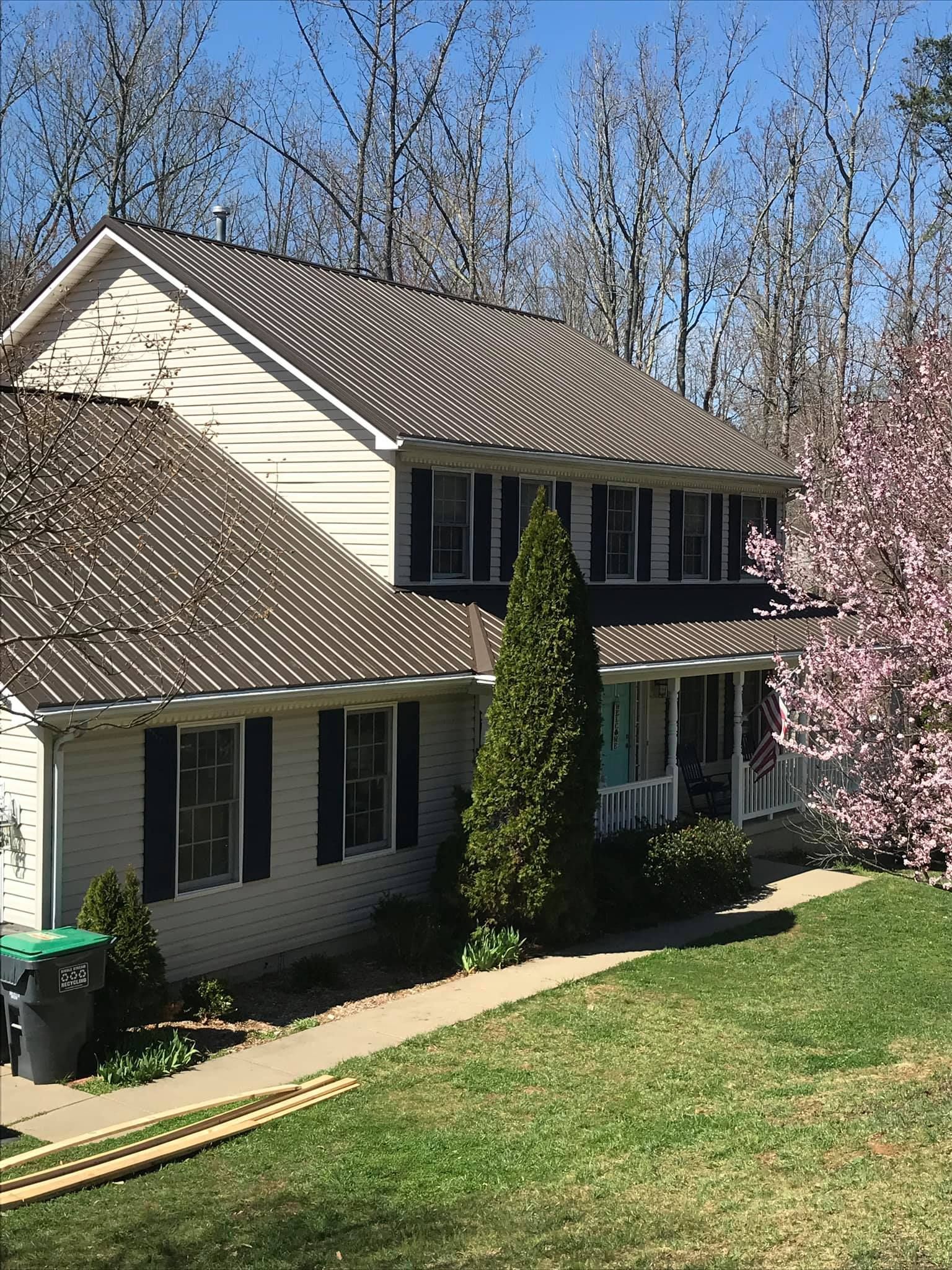 A white house with a brown metal roof and black shutters is surrounded by trees.