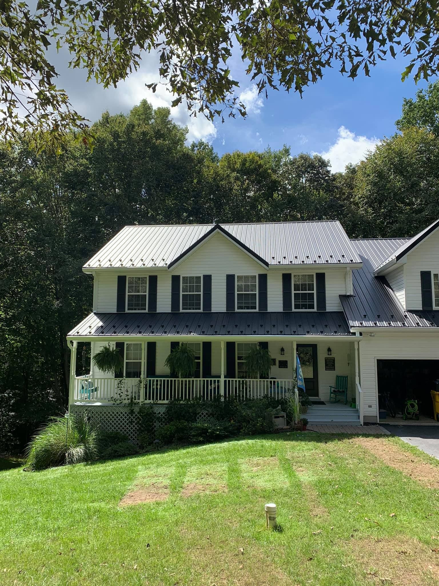 A white house with a gray metal roof and black shutters is surrounded by trees.