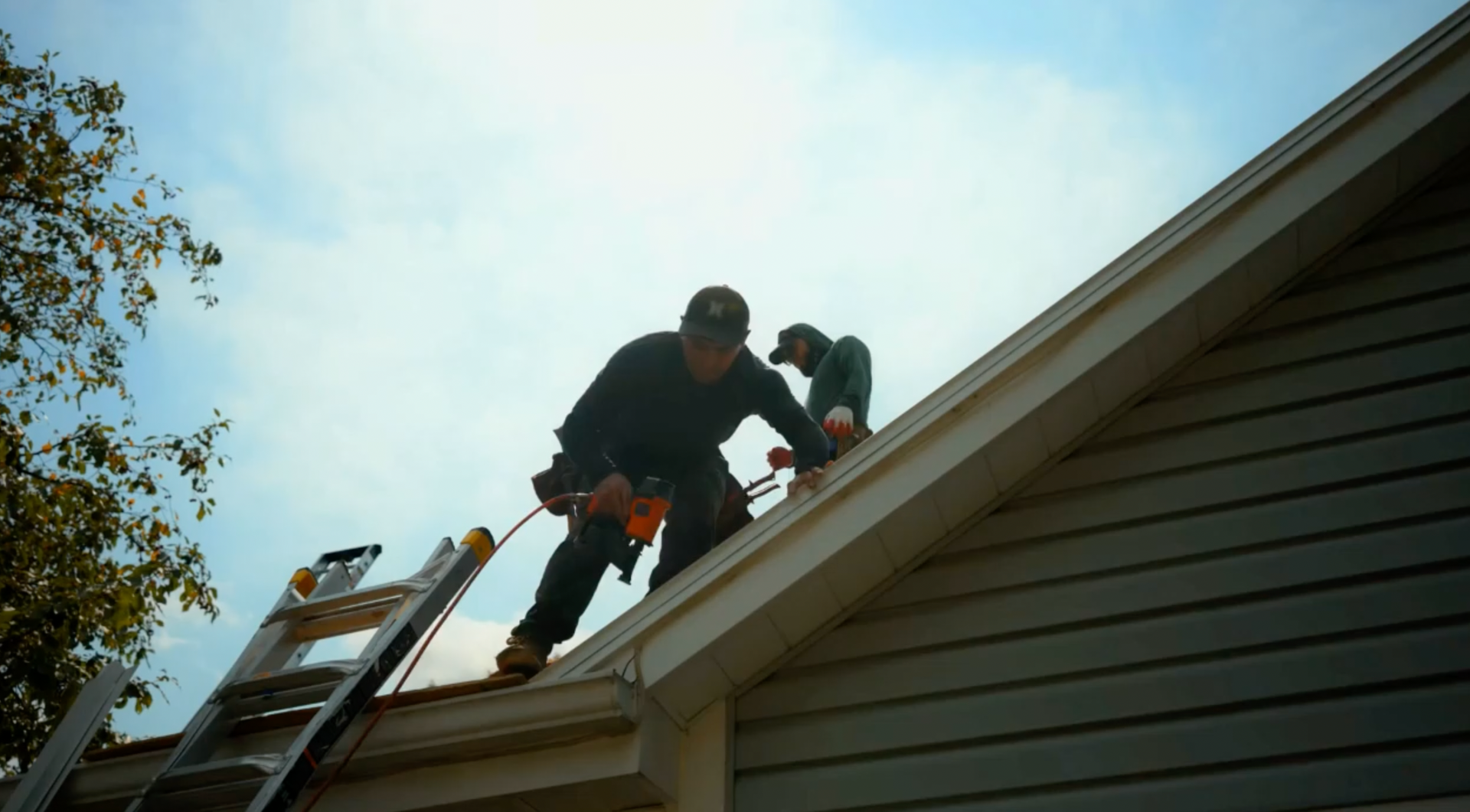 Two roofers working on a house roof, one using a tool, ladder beside the house, blue sky background.
