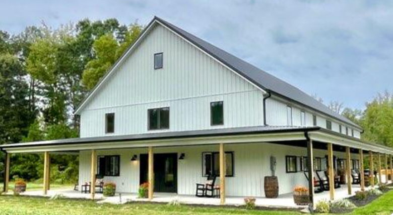 White barn-style farmhouse with a wraparound porch and green trim, set in a grassy rural landscape.