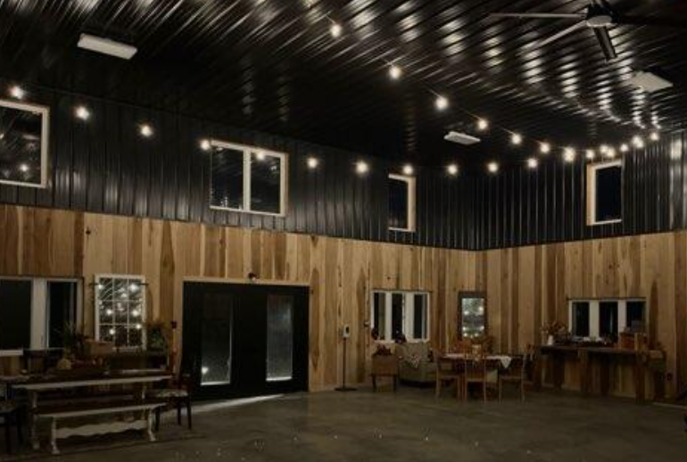 Inside a barn-like venue, wooden walls, black ceiling, string lights, large black doors, and windows.