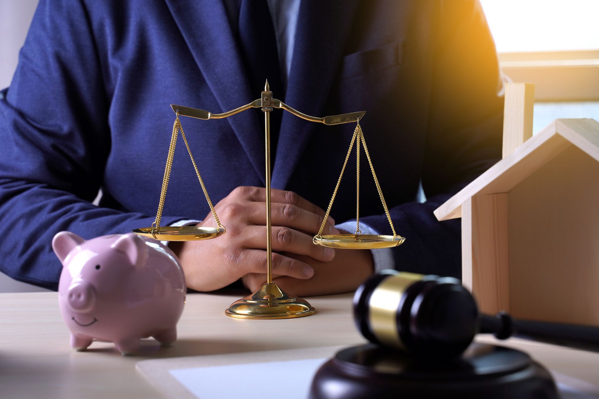 Person behind scales, gavel, piggy bank, and house model on a table, possibly legal or financial context.
