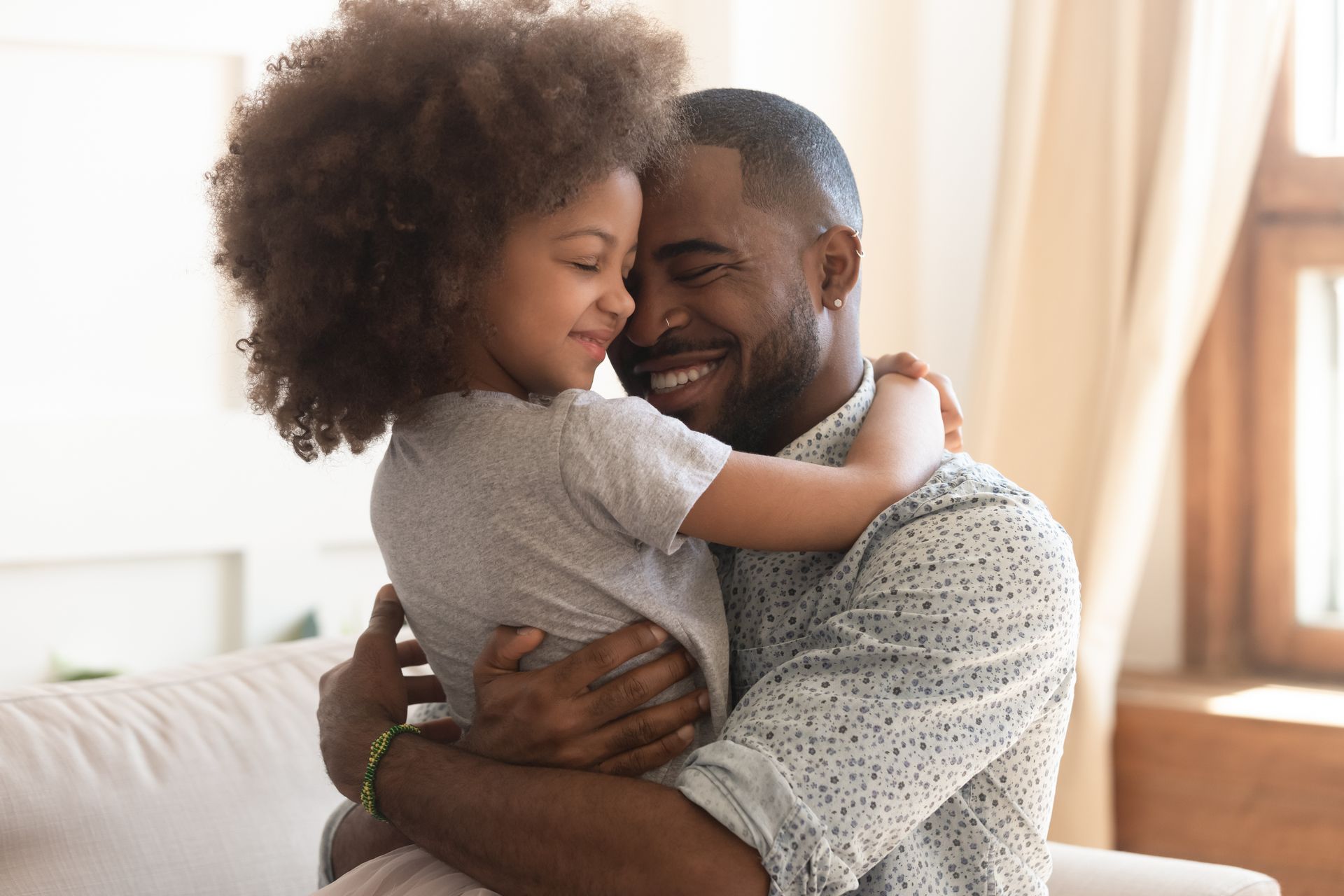 A smiling person embracing a child. Both appear happy indoors, near a window.