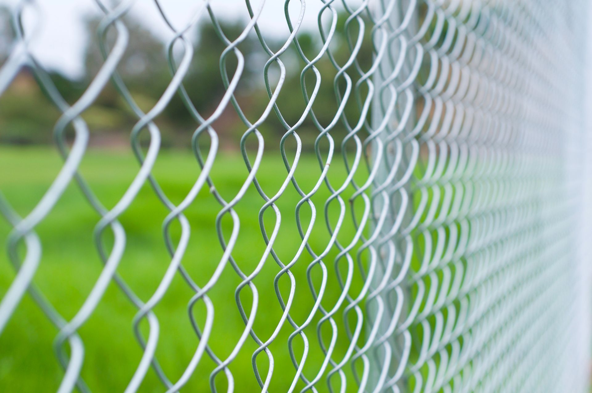 Close-up of a galvanized chain-link fence with a blurred green grassy field behind it