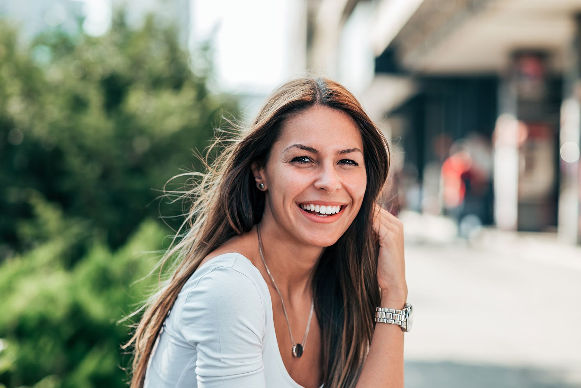 Woman with long brown hair smiles outdoors, wearing white shirt and jewelry.