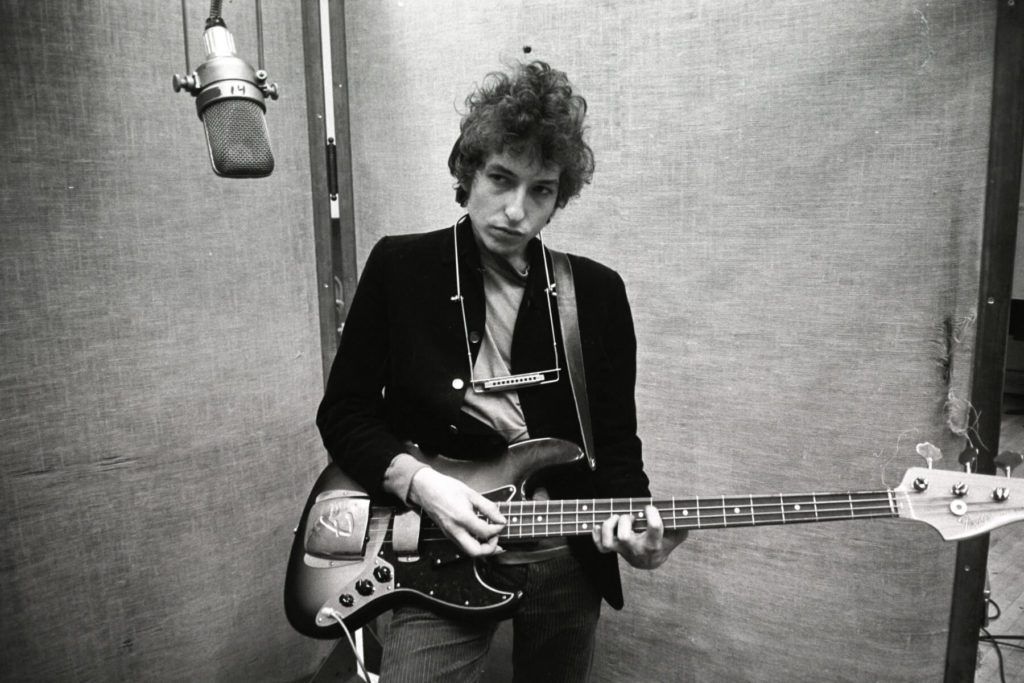 Bob Dylan playing a bass guitar in a recording studio. He's wearing a jacket and looking down at the instrument. A microphone hangs above.