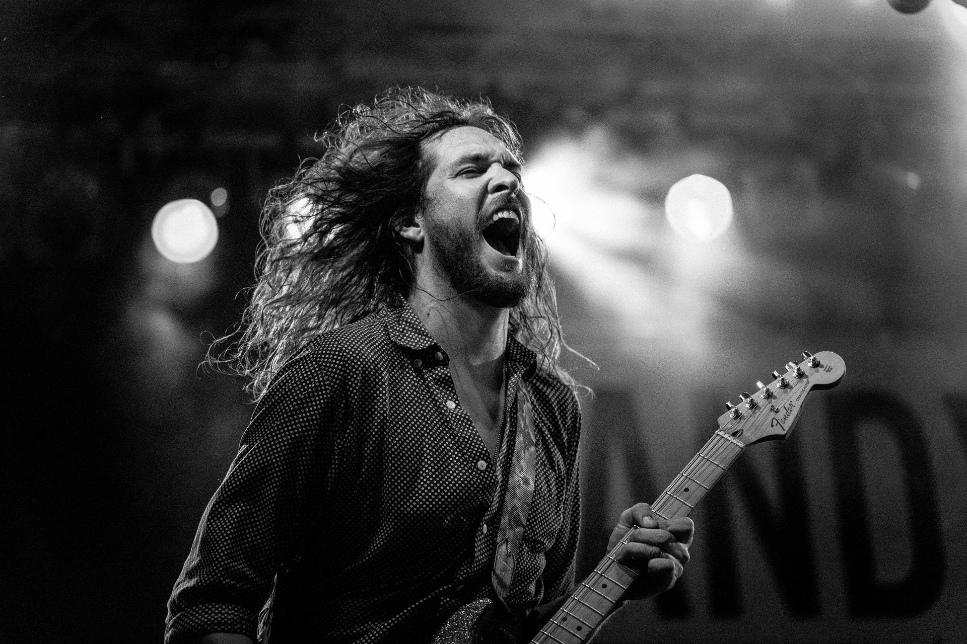 Man with long hair playing electric guitar and singing loudly on stage, mouth wide open.