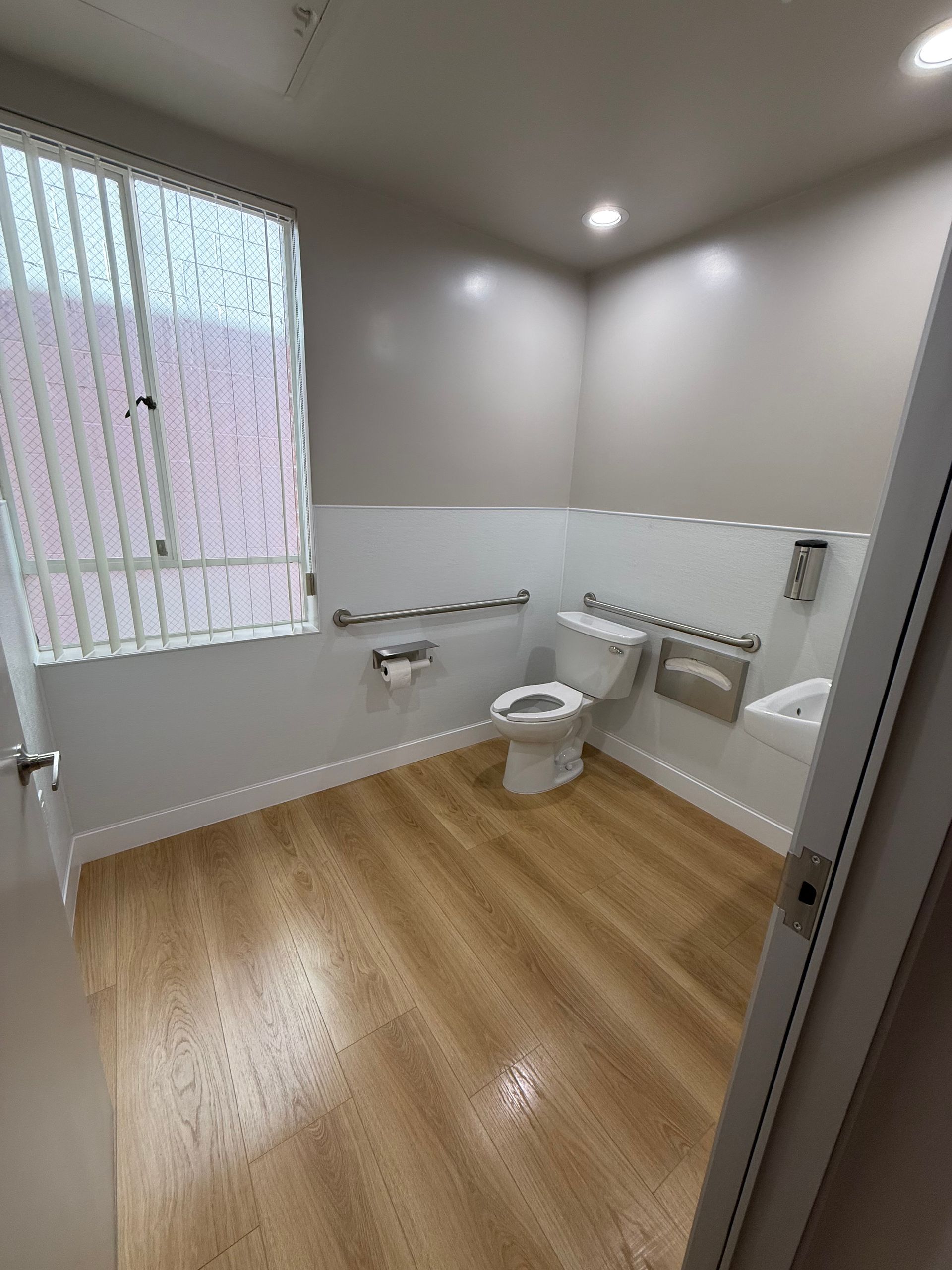 Small accessible bathroom with wood-look floor, white walls, toilet, sink, grab bars, and frosted window.