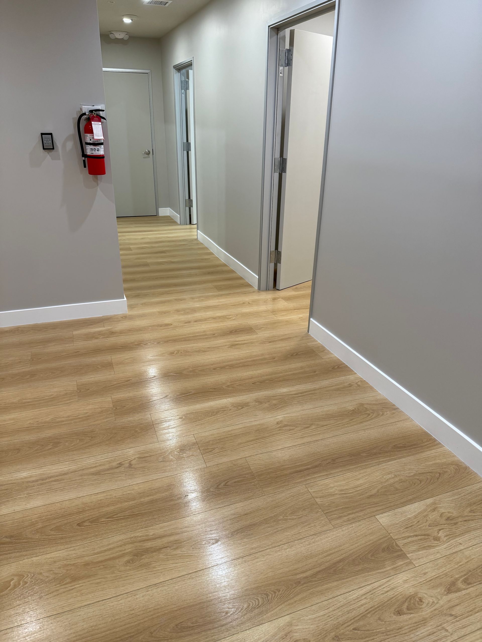 Empty office hallway with light wood flooring, gray walls, open doorways, and a red fire extinguisher on the left.
