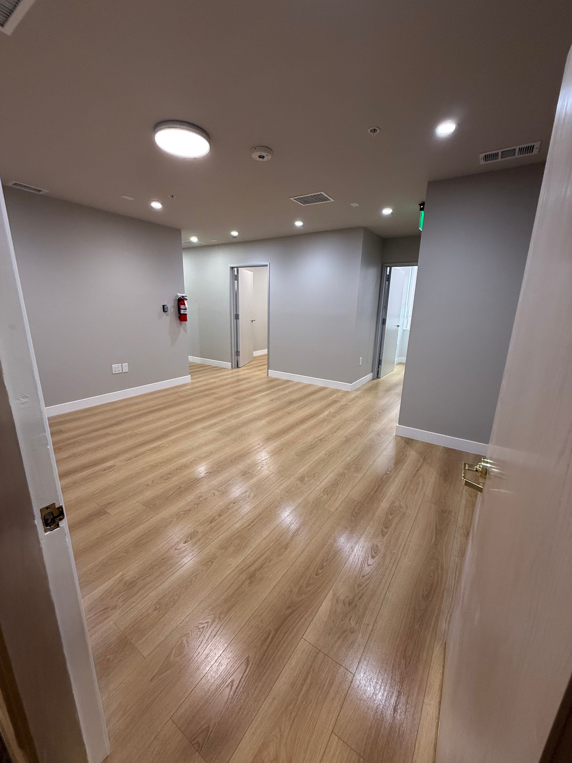 Empty modern room with light wood floors, gray walls, recessed lights, and a door at the back.
