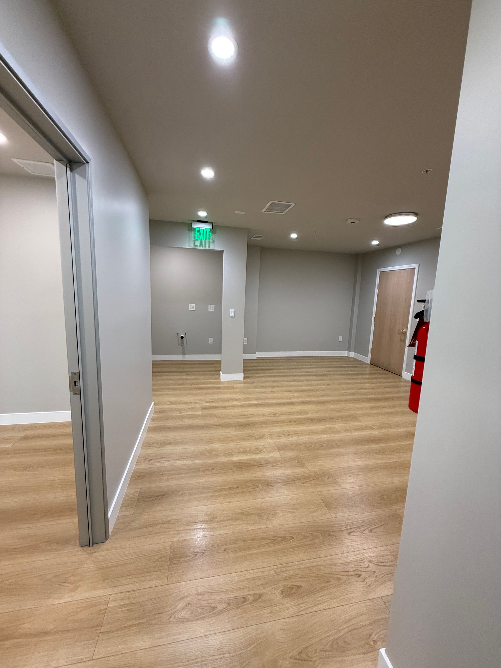 Empty office hallway with wood flooring, gray walls, exit sign, and fire extinguisher on the right.