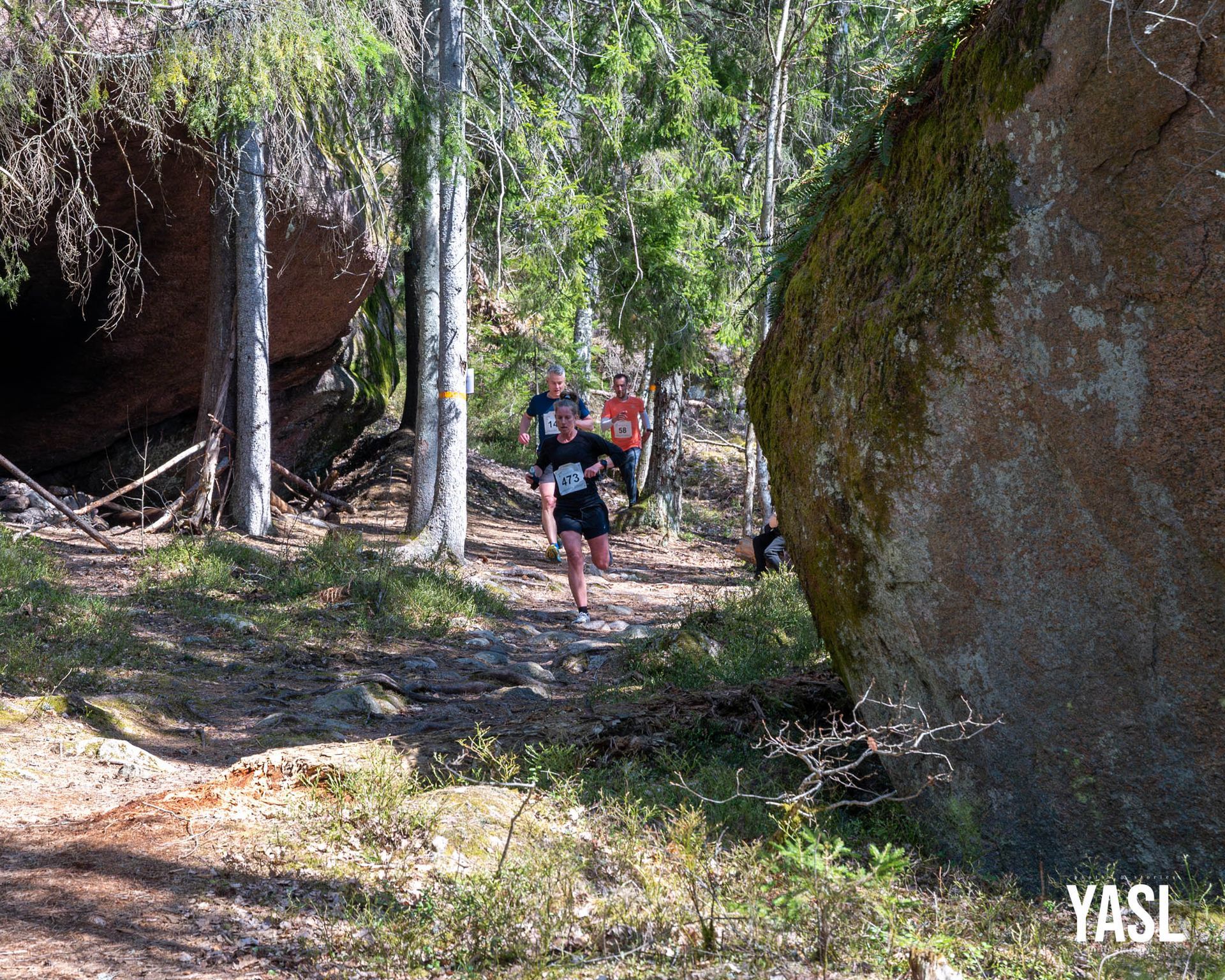 A group of people are walking through a forest next to a large rock.