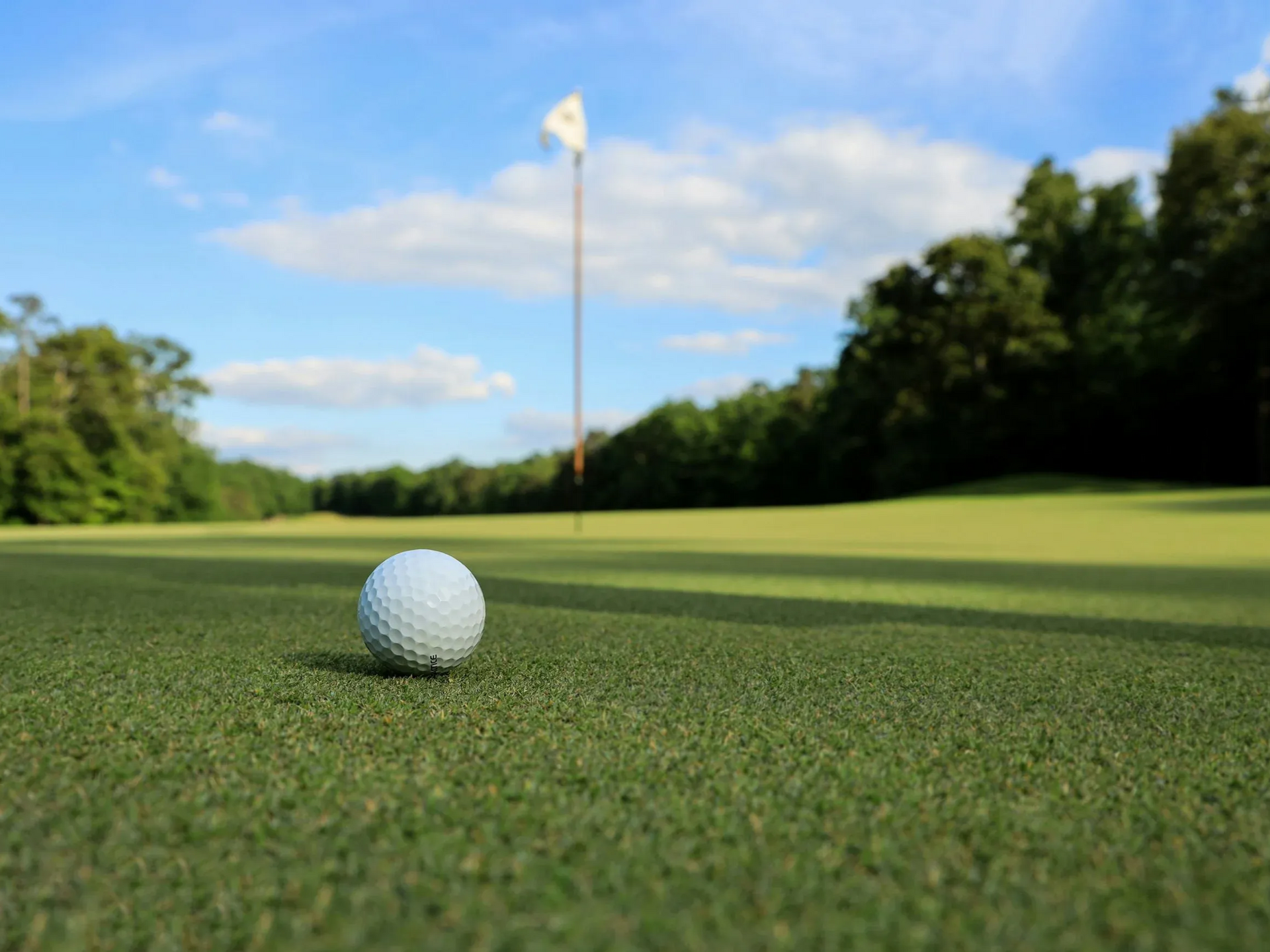 Golfball auf dem Grün nahe dem Loch, im Hintergrund Bäume und blauer Himmel.