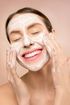 A woman is smiling while washing her face with a white mask.
