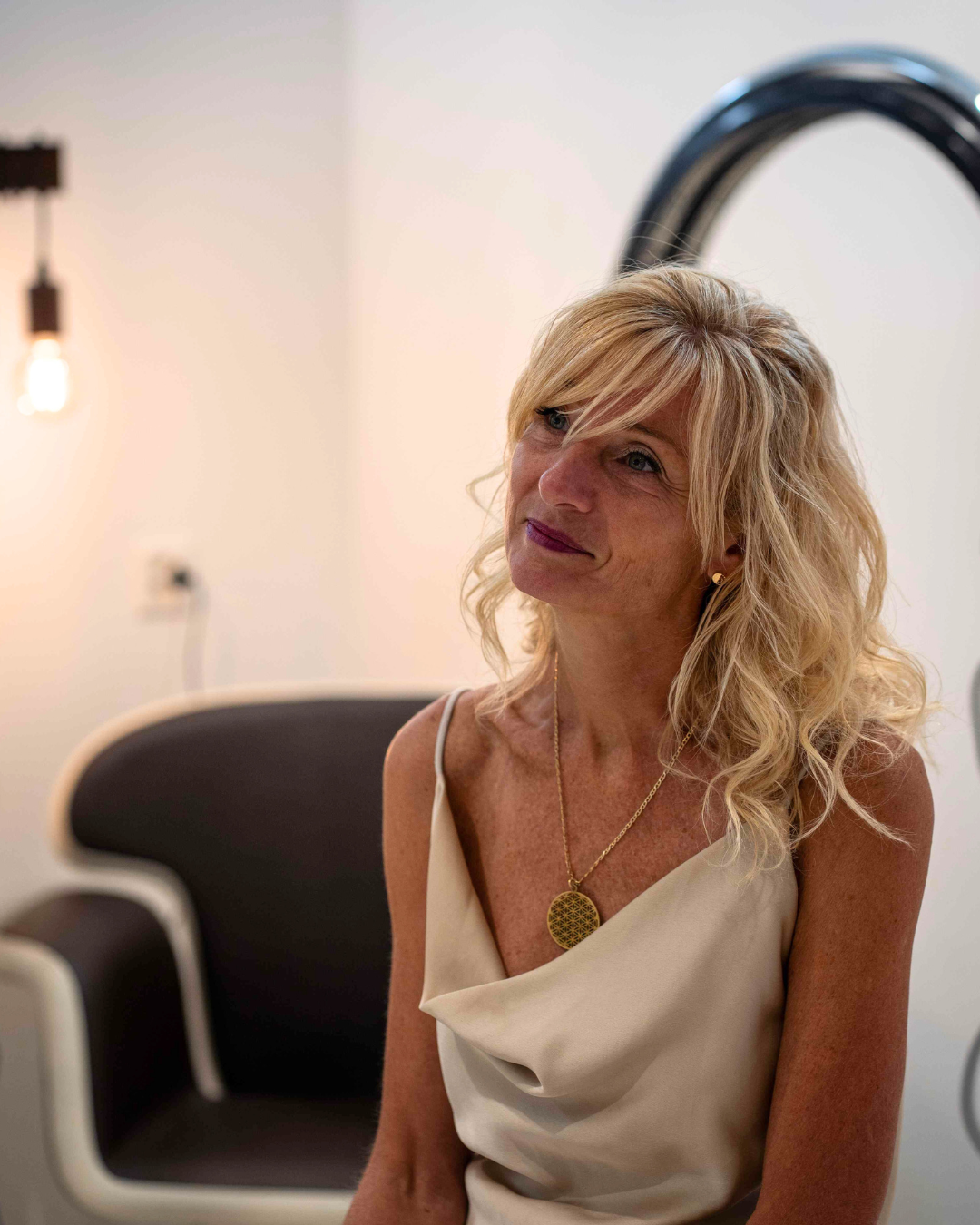Photo-Laurence Femme vêtue d'un haut couleur crème, regardant vers le haut, portant un collier pendentif, assise près d'une chaise noire et blanche.
