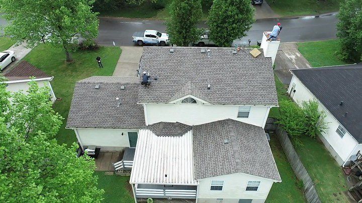 Aerial view of a house with a gray shingled roof, surrounded by trees and parked cars nearby