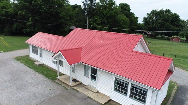 White building with red metal roof beside a gravel lot and green field, viewed from above
