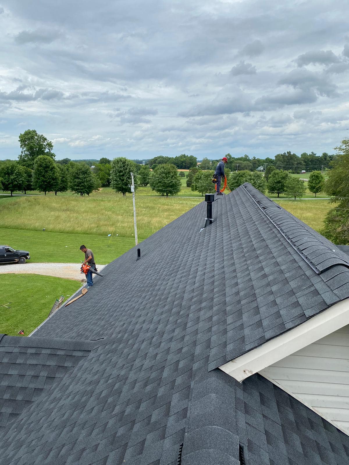 Two workers on a dark gray shingle roof, one near the peak and one at the edge, in a grassy rural area.