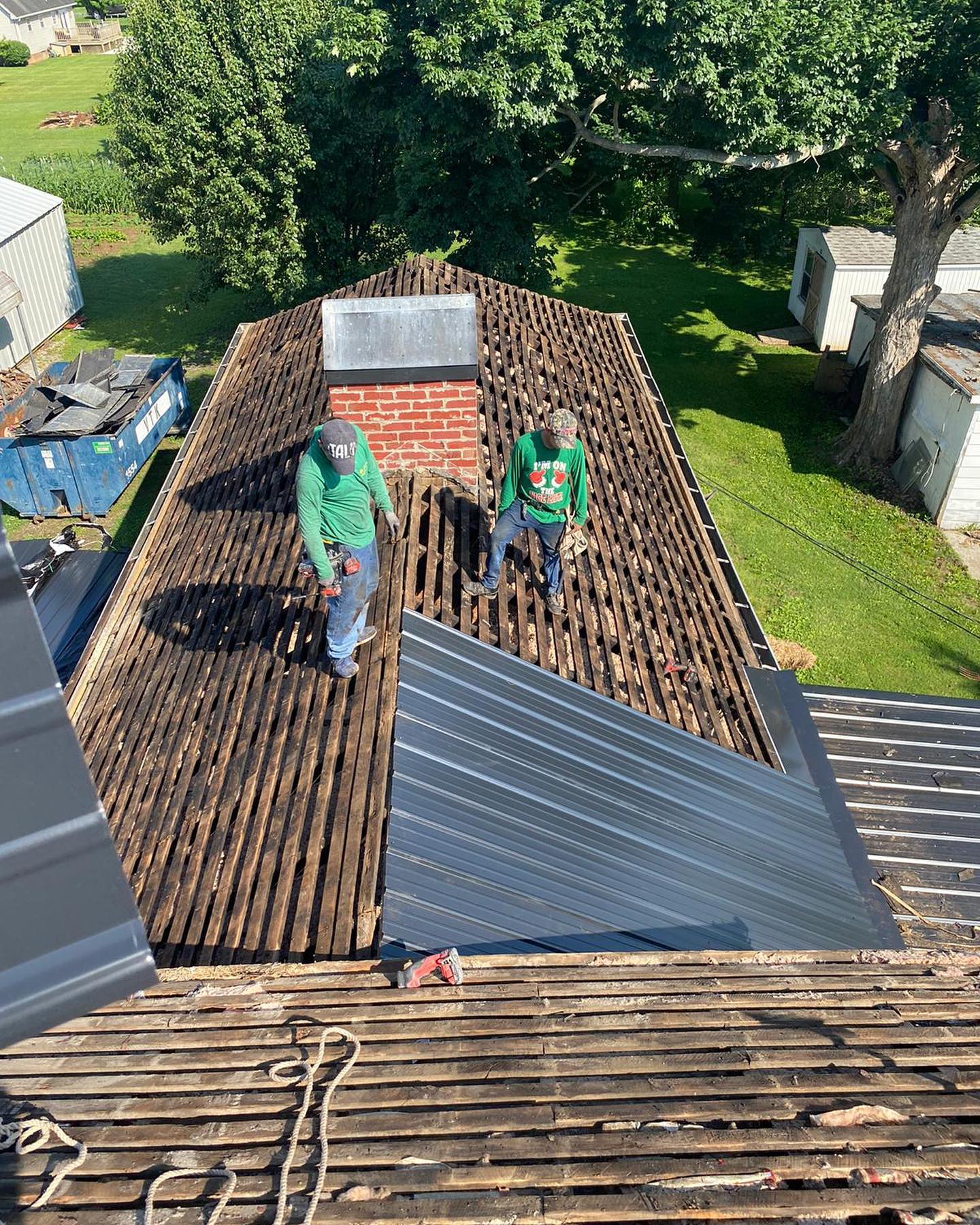 Workers installing metal roofing on a house roof, with exposed wooden sections and trees in the background