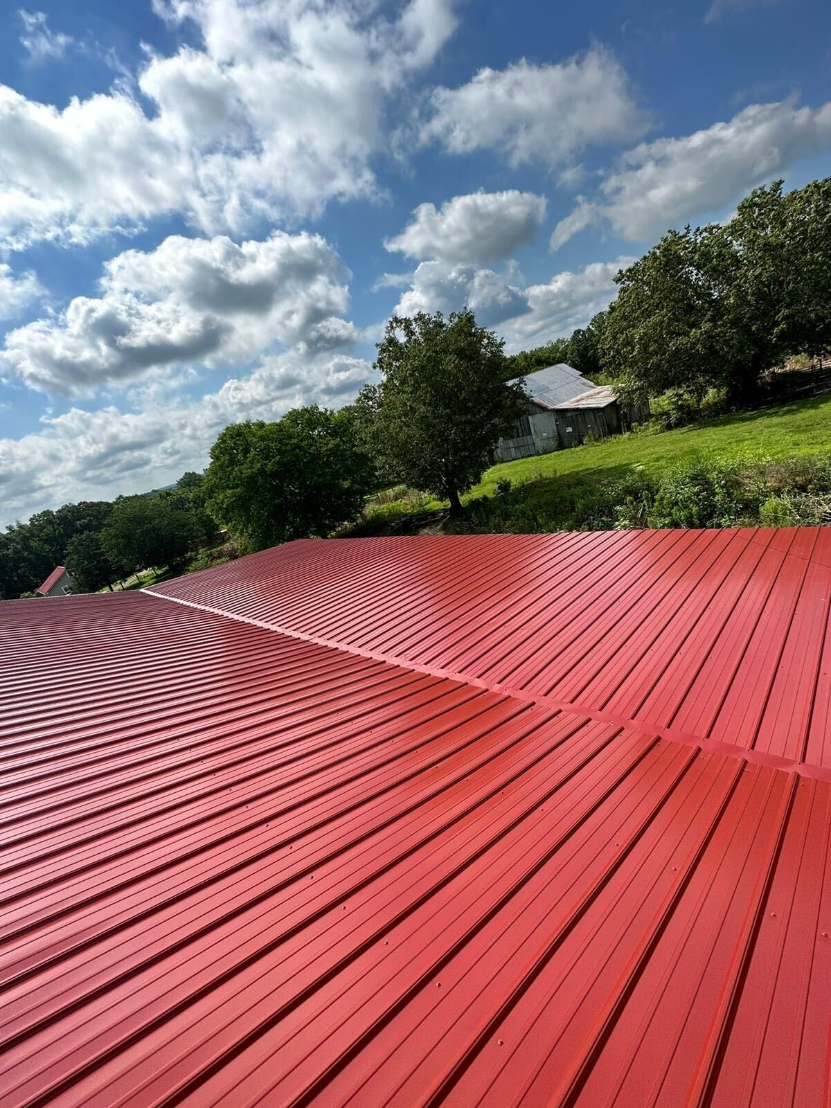Red corrugated metal roof in the foreground with green trees, grassy hill, and cloudy blue sky beyond