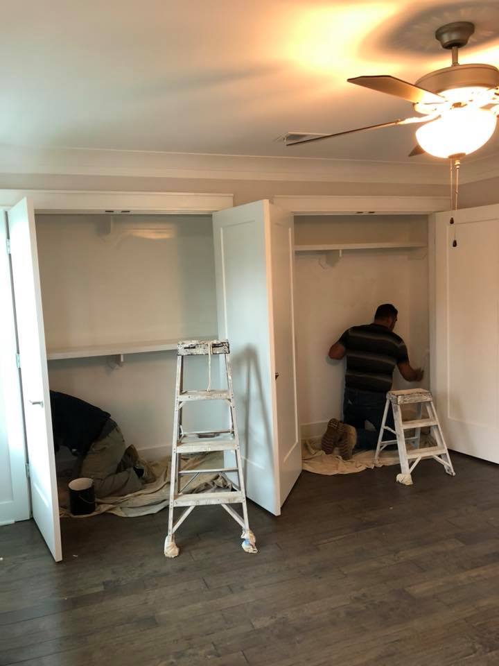 Two workers painting white closets in a bedroom with two step ladders and dark wood flooring.