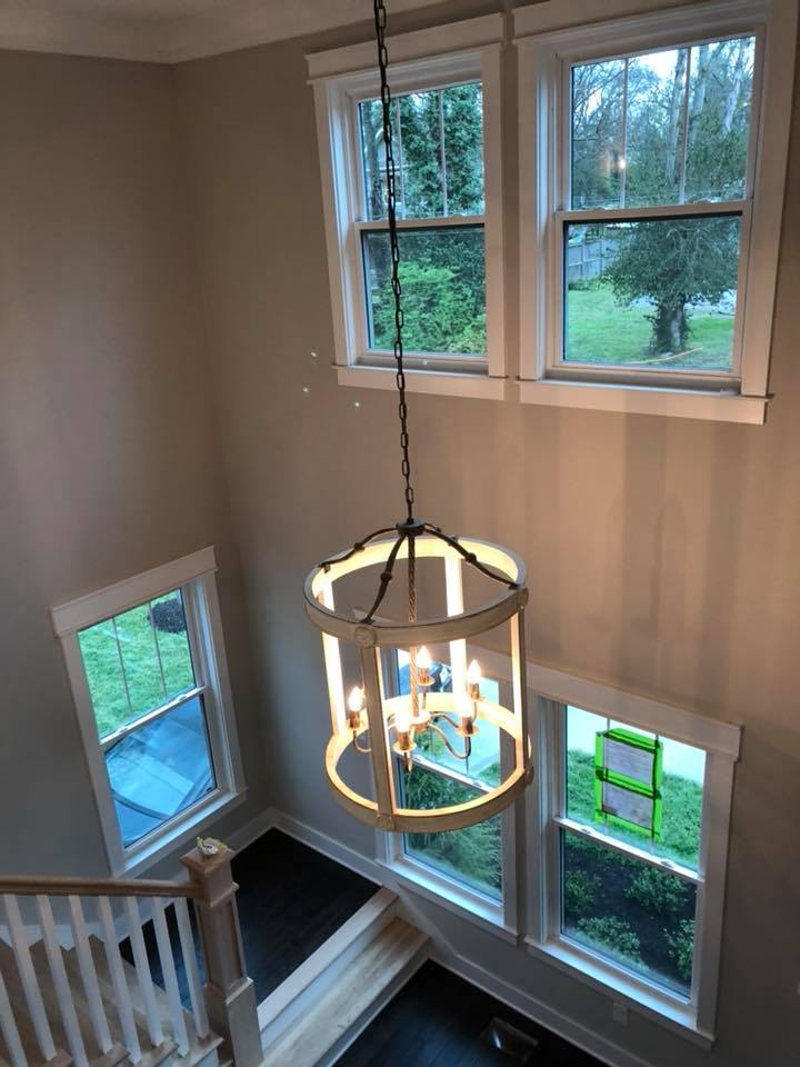 Stairwell with hanging chandelier, white railings, and large windows overlooking green trees.