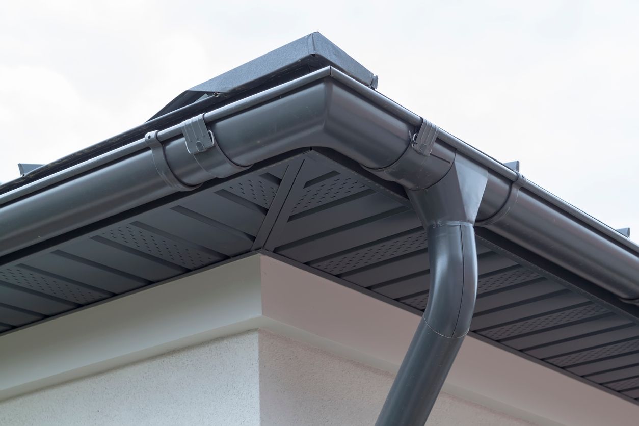 Corner of a house roof with gray gutters and a downspout against a cloudy sky