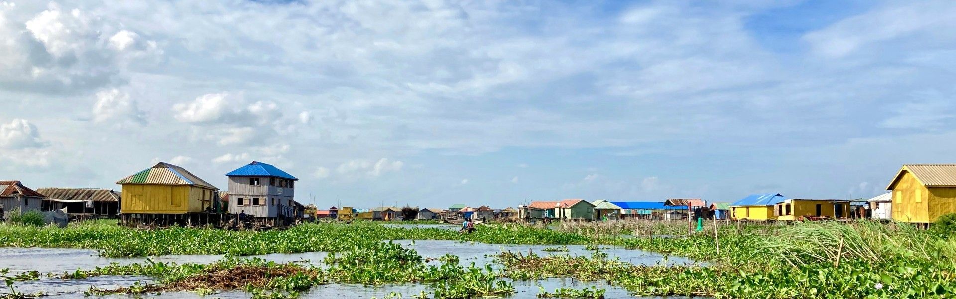 This scenic image captures the unique beauty of Ganvié, Benin’s remarkable lake village built on stilts above the waters of Lake Nokoué. Colorful wooden homes, narrow waterways, and lush floating vegetation create an extraordinary landscape where daily life has flourished on the water for centuries. Often called the