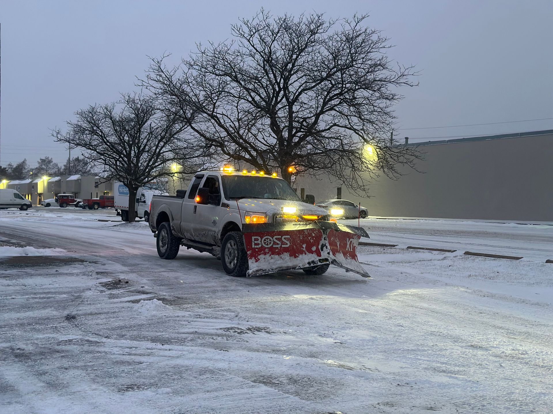 A white pickup truck with a red snowplow attached to the front drives through a snow-covered parking lot at dusk.