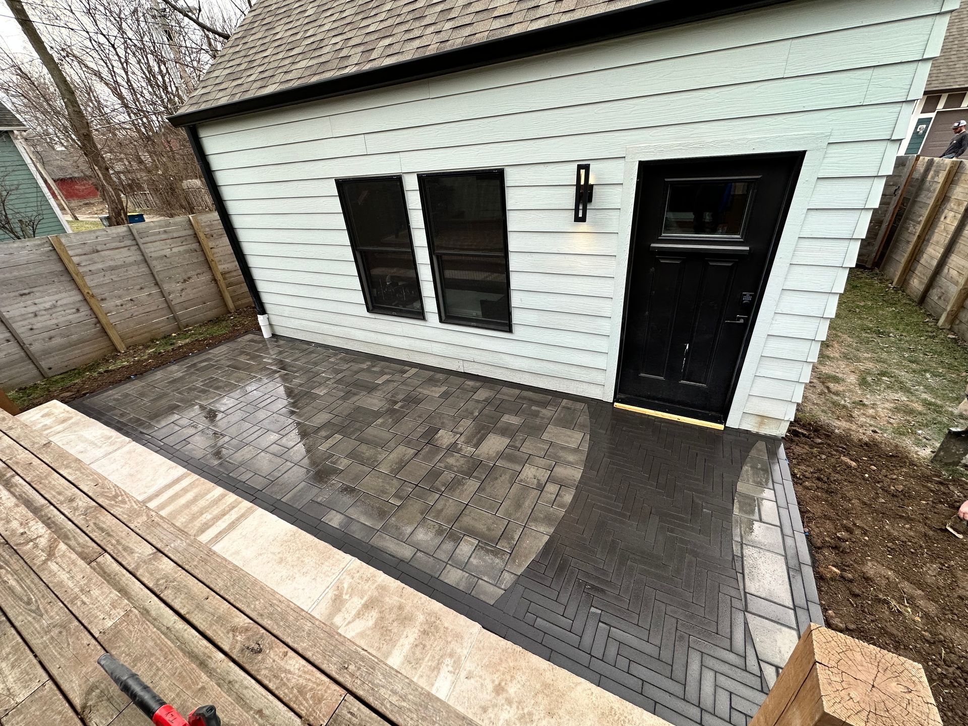 A light-colored building with a black door and windows, featuring a new gray stone paver patio and a wooden deck.