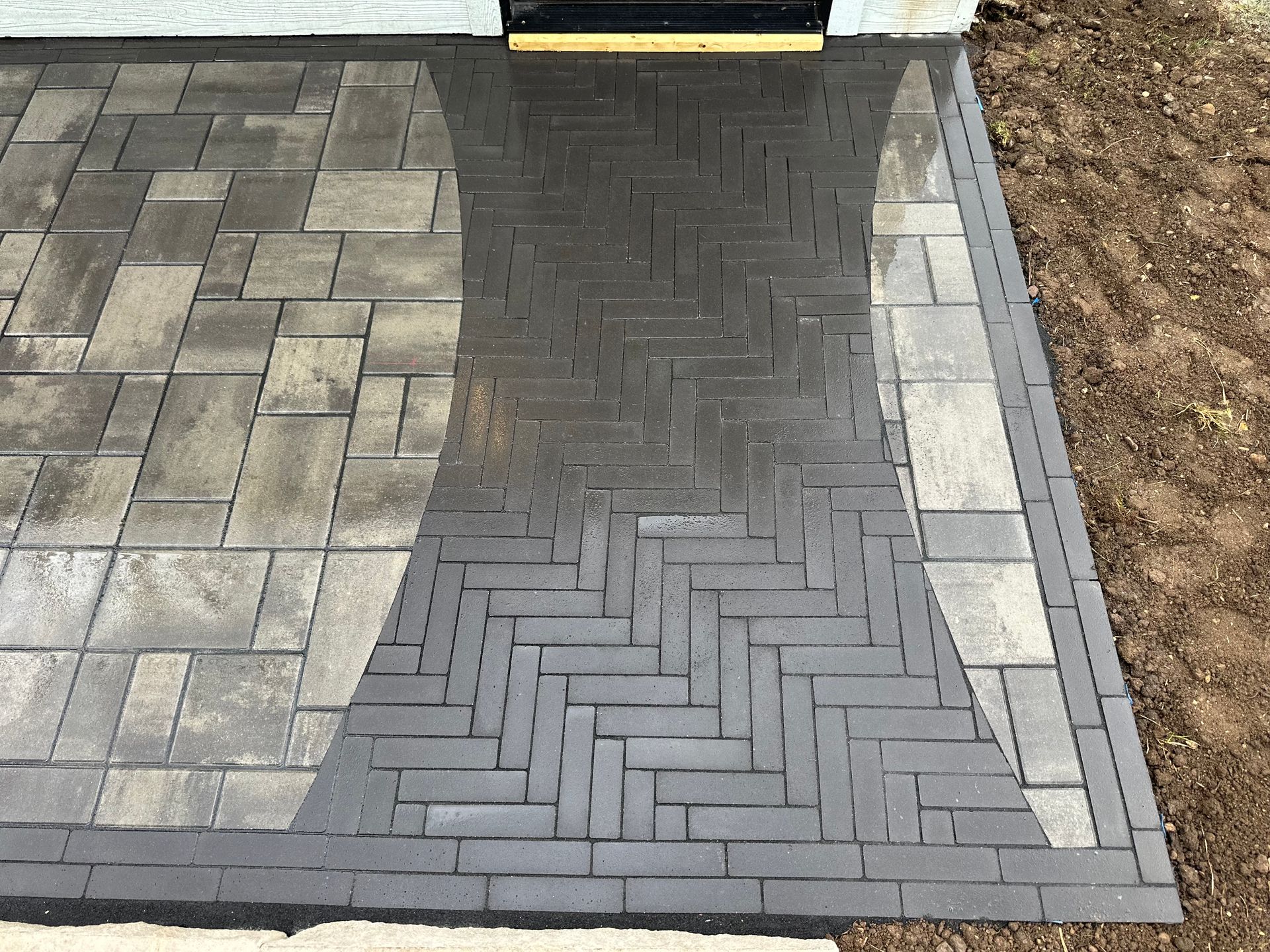 A brick paver walkway leading to a doorway, featuring a dark herringbone-patterned path bordered by lighter rectangular tiles.