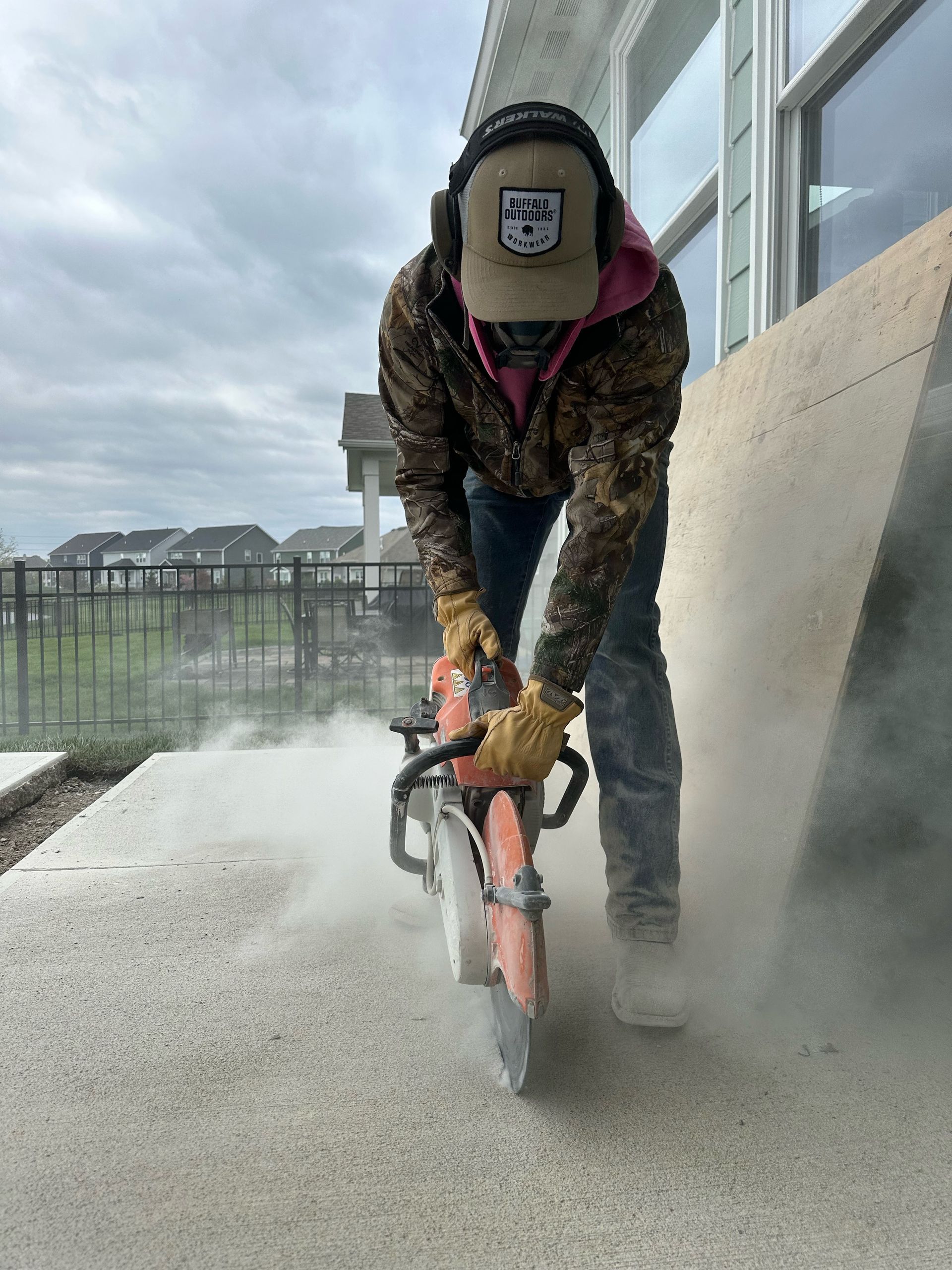 A person wearing protective gear cuts into a concrete surface with a powered saw, creating a cloud of dust.