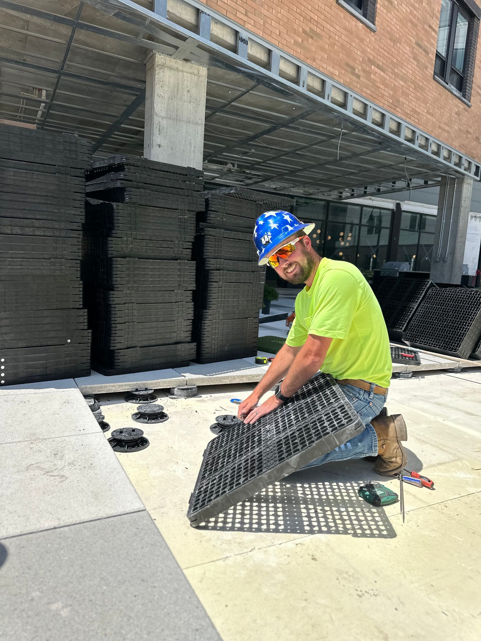 A construction worker in a high-vis shirt and hard hat installs black modular paving tiles on a patio pedestal system.