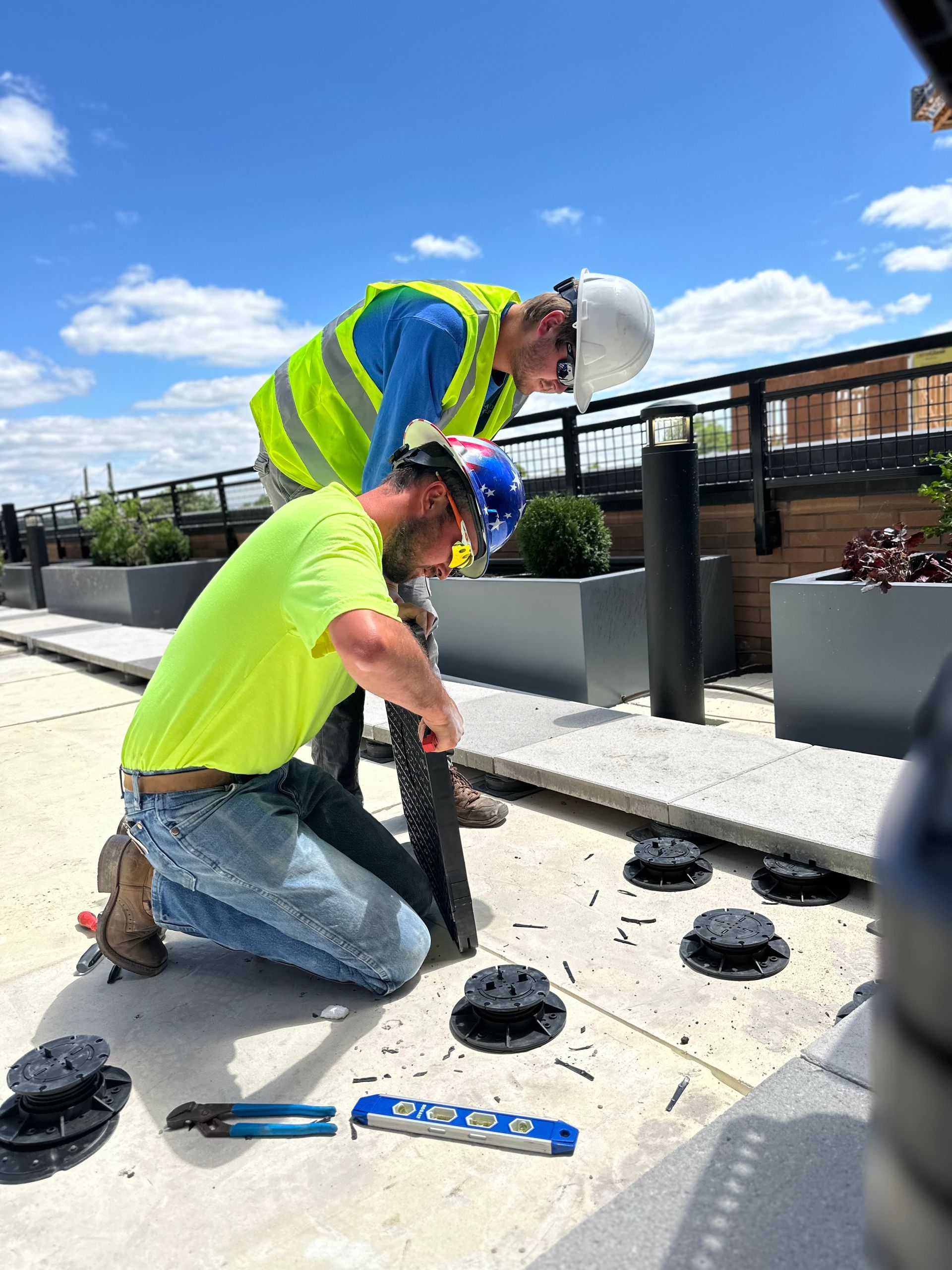 Two construction workers in high-visibility vests install adjustable pedestal supports for pavers on a sunny rooftop terrace.