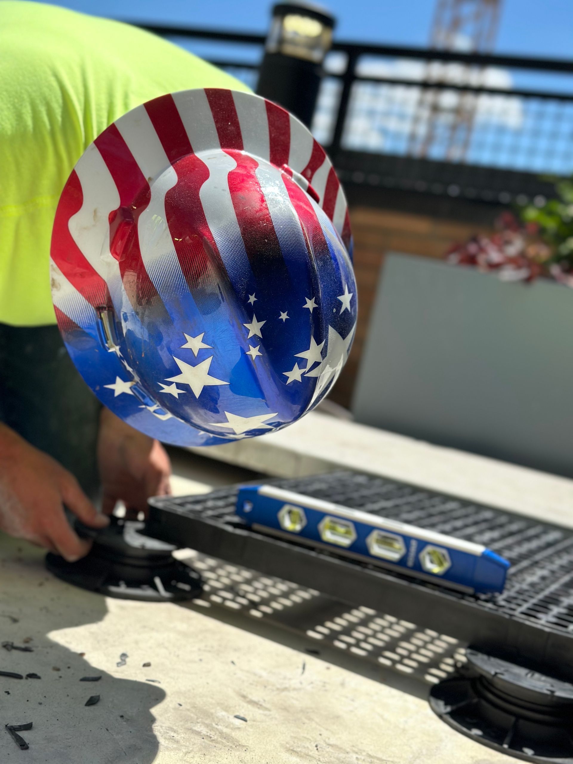 A construction worker wearing an American flag hard hat uses a spirit level on a metal grate on a rooftop.