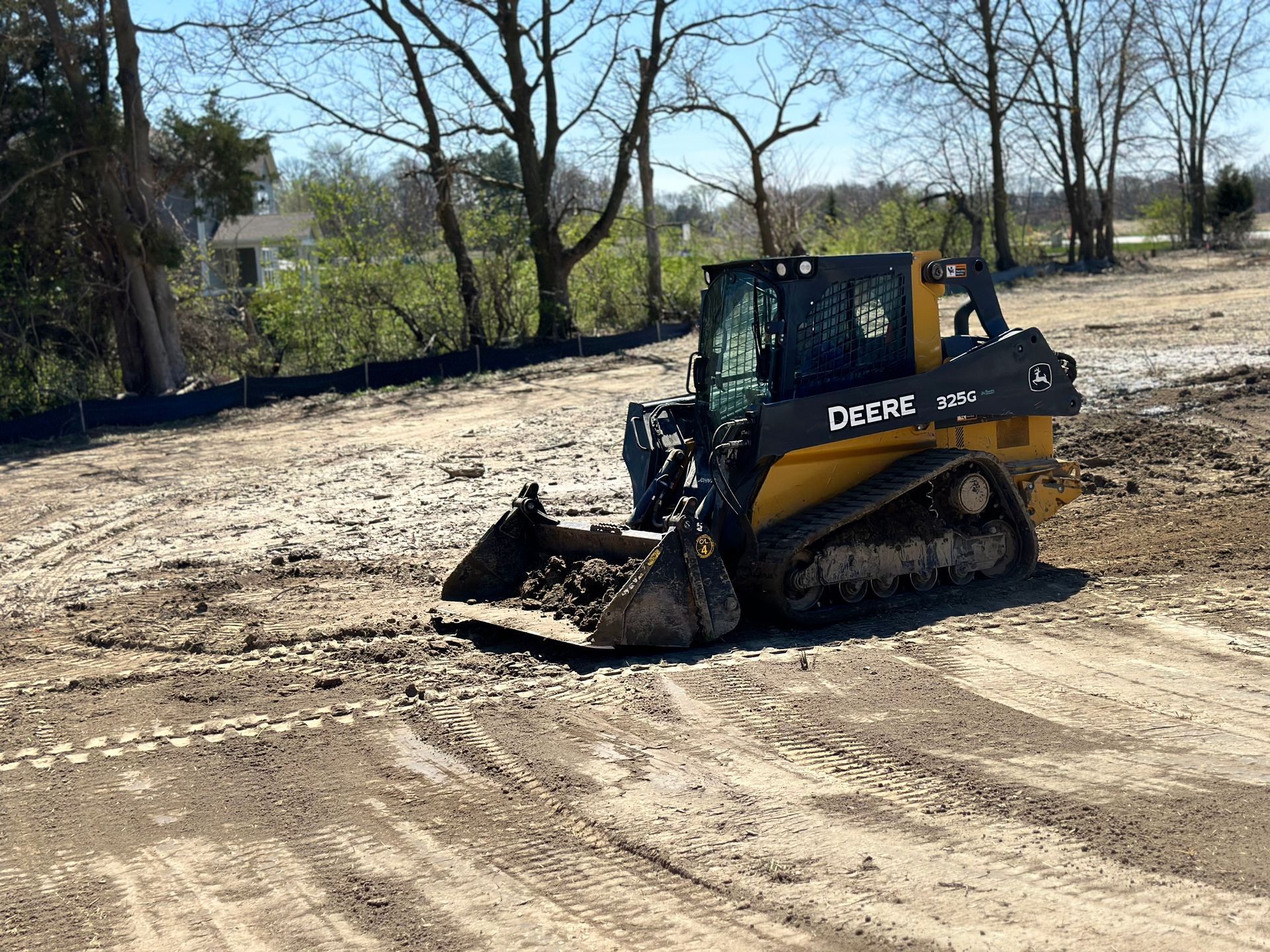 A yellow John Deere compact track loader moves dirt on a sunny, unpaved construction site near a tree line.