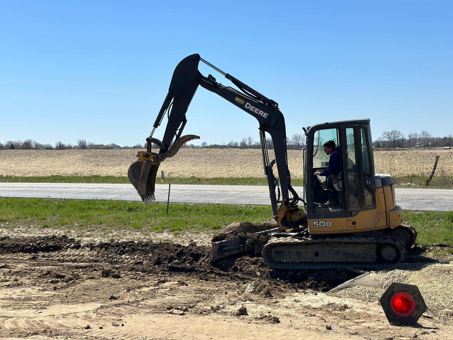 A yellow excavator with its bucket raised, operating in a field near a road under a clear blue sky.