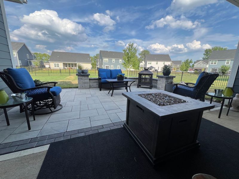 A backyard stone patio with blue cushioned chairs, a fire pit, and a black outdoor rug under a blue, cloudy sky.
