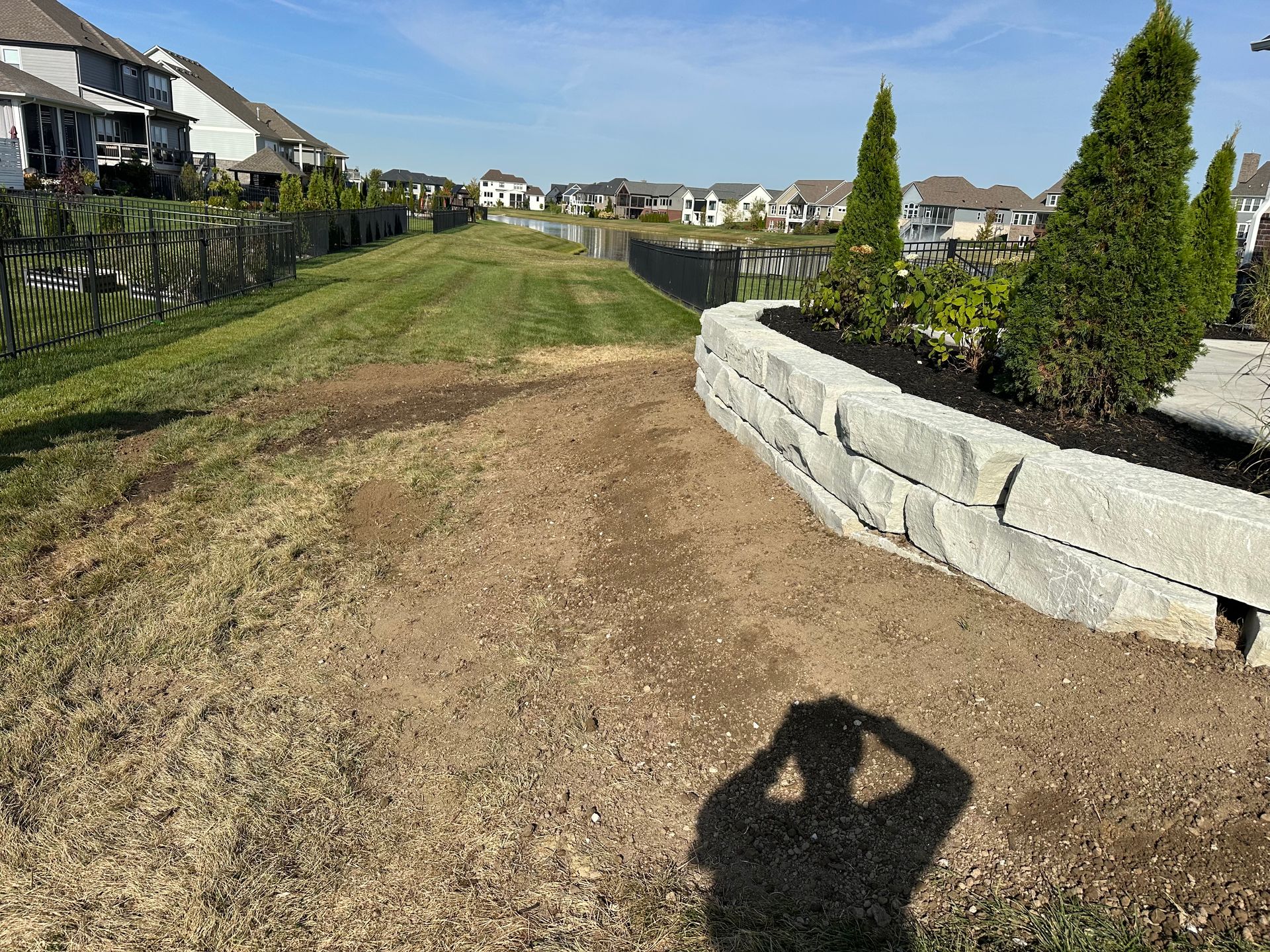 A stone retaining wall with evergreens in a residential yard, featuring a grassy strip and a person's shadow in the front.